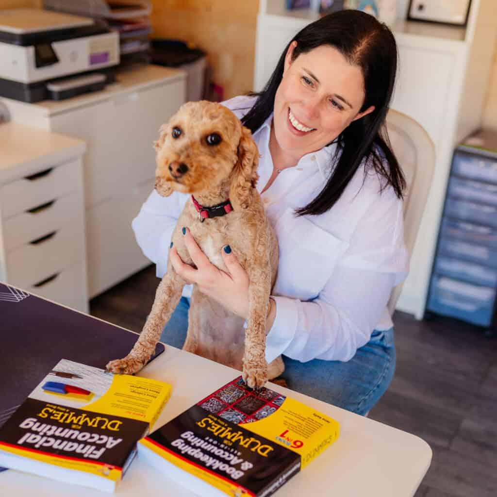 Woman smiling with small dog at office desk with accounting books.